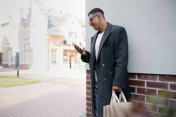 Homme afro-américain joyeux en manteau et lunettes de soleil tenant des sacs à provisions et bavardant sur le téléphone portable près du bâtiment — Photo de stock