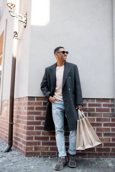 Longitud completa de hombre afroamericano con estilo en abrigo y gafas de sol de pie con la mano en el bolsillo de jeans y bolsas de compras - foto de stock