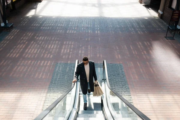 Vista de ángulo alto del hombre afroamericano de moda con bolsas de compras en escaleras mecánicas - foto de stock