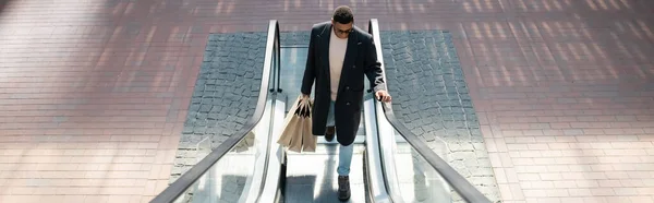 Vista de ángulo alto del hombre afroamericano de moda con bolsas de compras en escaleras mecánicas, pancarta - foto de stock