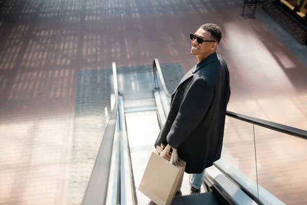 Hombre afroamericano de moda en gafas de sol y abrigo sonriendo con bolsas de compras en escaleras mecánicas - foto de stock