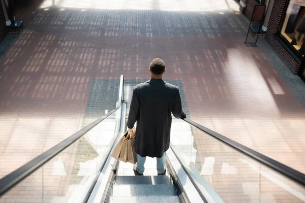 Vista trasera del hombre afroamericano en abrigo de pie con bolsas de compras en escaleras mecánicas - foto de stock