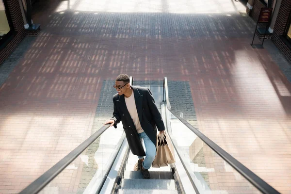 Visão de alto ângulo de sorrir e moderno homem americano africano com sacos de compras na escada rolante — Fotografia de Stock