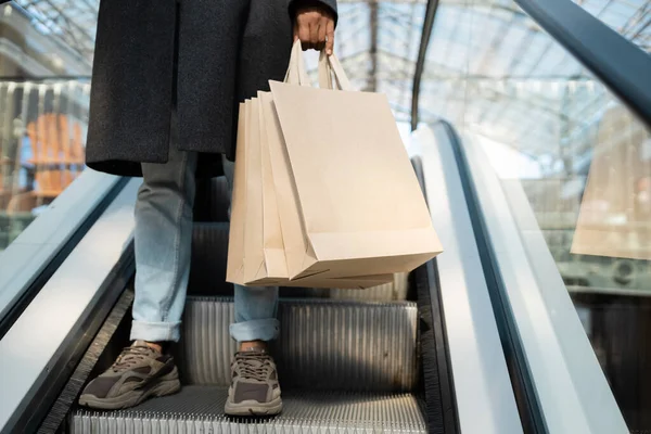 Vista recortada del hombre afroamericano en abrigo y zapatillas de deporte sosteniendo bolsas de compras en escaleras mecánicas - foto de stock