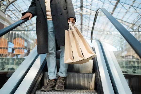 Vista parcial del hombre afroamericano en abrigo y zapatillas de deporte sosteniendo bolsas de compras en escaleras mecánicas - foto de stock