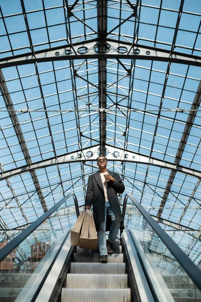 Vista de ángulo bajo de hombre afroamericano con estilo con bolsas de compras y taza de papel en escaleras mecánicas bajo techo de cristal - foto de stock