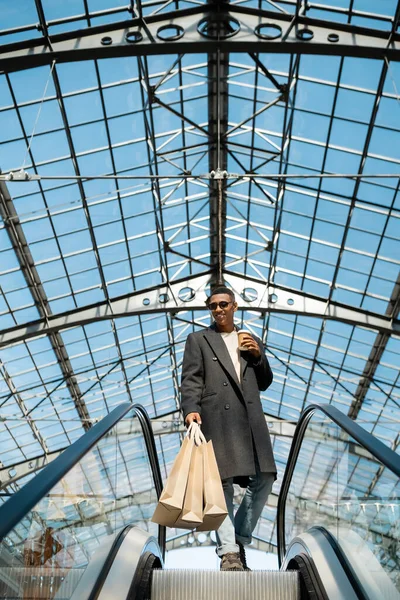 Vista basso angolo di uomo afro americano felice ed elegante con borse della spesa e drink da asporto sulla scala mobile sotto il tetto trasparente — Foto stock