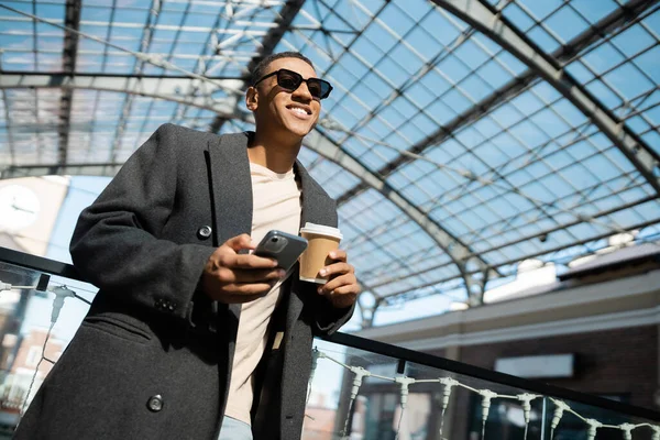 Низький кут зору на модного афроамериканця з смартфоном і кавою, щоб посміхатися під скляним дахом на відкритому повітрі — стокове фото