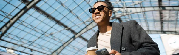 Visão de baixo ângulo do homem americano africano na moda com copo de papel e celular sob telhado de vidro, bandeira — Fotografia de Stock