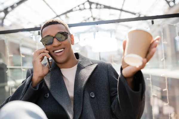 Cheerful african american man in sunglasses holding paper cup and talking on mobile phone outdoors — Stock Photo