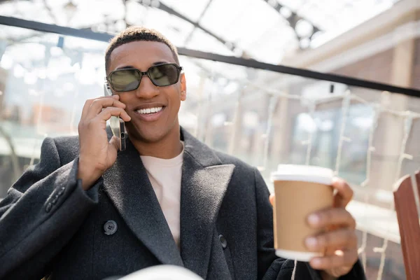 Hombre afroamericano de moda con café para ir sonriendo durante la conversación en el teléfono inteligente - foto de stock