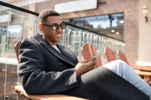 Smiling african american man in trendy coat and sunglasses chatting on mobile phone while sitting on city street — Stock Photo