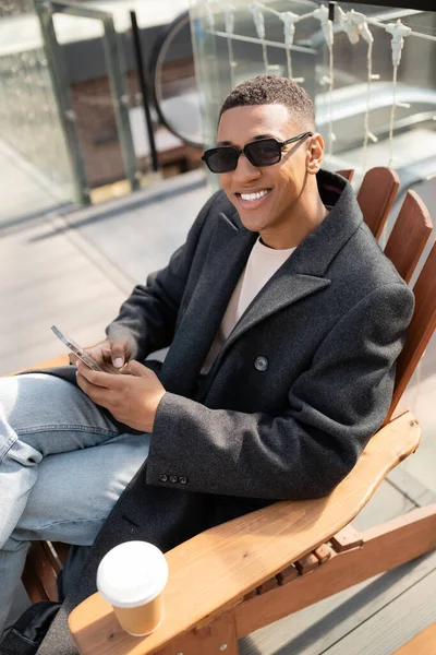 Uomo afroamericano in cappotto e occhiali da sole seduto con il telefono cellulare e sorridente alla fotocamera sulla strada urbana — Foto stock