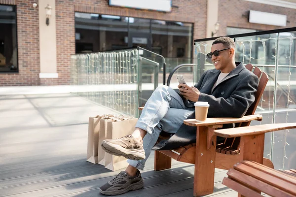 Uomo afroamericano alla moda in occhiali da sole seduto con smartphone vicino tazza di carta e borse della spesa — Foto stock