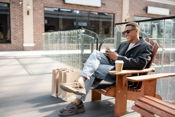 Trendy african american man in sunglasses sitting with smartphone near paper cup and shopping bags — Stock Photo