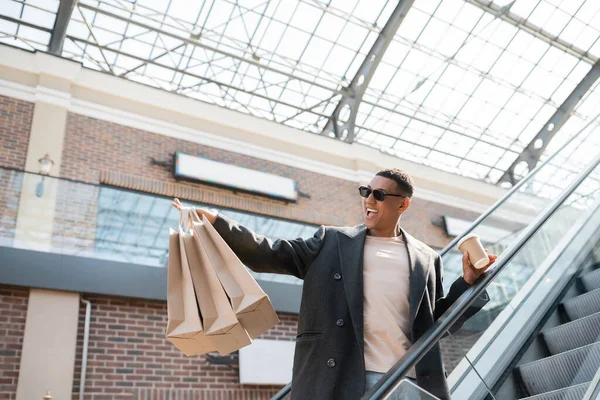 Excited african american man in trendy coat and sunglasses holding coffee to go and shopping bags on escalator — Stock Photo