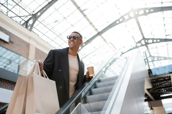 Alegre afroamericano hombre en gafas de sol mirando hacia otro lado en escaleras mecánicas mientras sostiene bolsas de compras y taza de papel - foto de stock