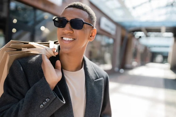 Hombre afroamericano feliz en abrigo y gafas de sol sosteniendo bolsas de compras en la calle borrosa - foto de stock