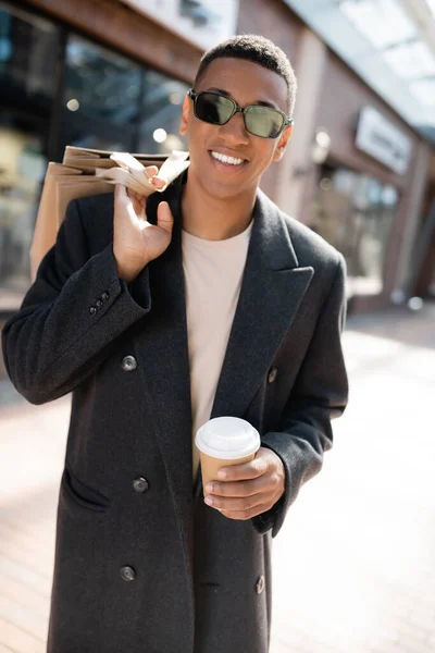 Hombre afroamericano de moda en gafas de sol con bebidas para llevar y bolsas de compras mientras sonríe a la cámara en la calle urbana - foto de stock