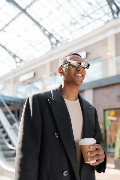 Стильный африканский американец в пальто и солнцезащитных очках с напитками на вынос и улыбкой на размытой улице — стоковое фото