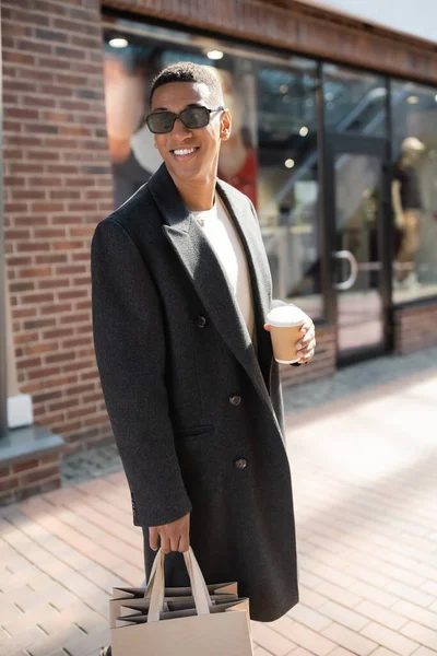 Trendy african american man with disposable cup and shopping bags smiling on urban street — Stock Photo