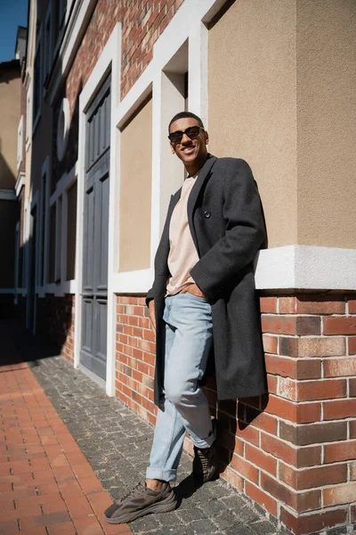 Longitud completa de hombre afroamericano en abrigo y gafas de sol de pie con la mano en el bolsillo de los vaqueros cerca de la construcción - foto de stock