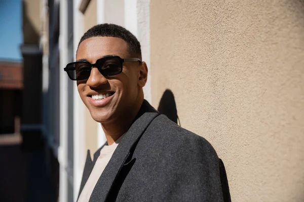 Heureux homme afro-américain en manteau et des lunettes de soleil élégantes regardant caméra près du mur — Photo de stock