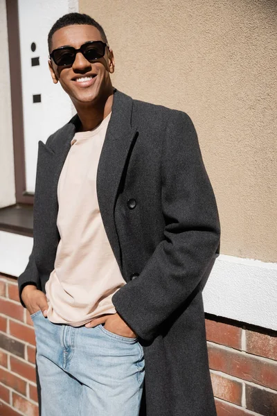 Homme afro-américain à la mode et heureux en lunettes de soleil et manteau debout avec les mains dans des poches de jeans près du bâtiment — Photo de stock