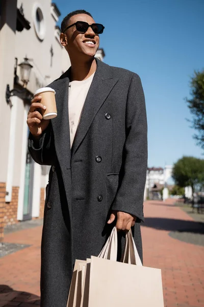 African american man in coat and sunglasses holding coffee to go and shopping bags while smiling on street — Stock Photo
