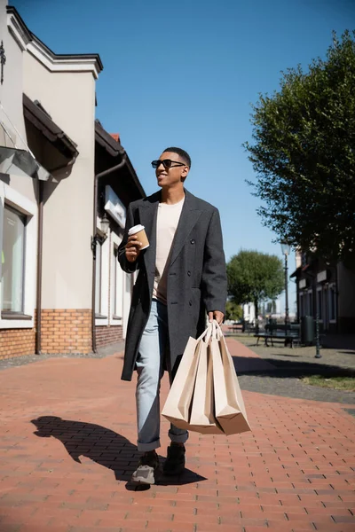 Longitud completa del hombre afroamericano de moda en gafas de sol caminando con bolsas de compras y taza de papel en la calle - foto de stock
