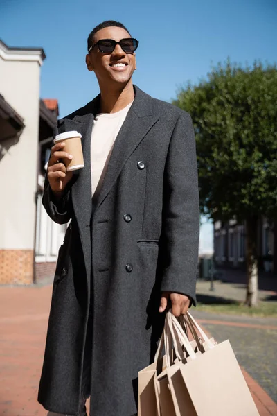 Щасливий афроамериканський чоловік у стильному пальто та сонцезахисні окуляри, що стоять з виносним напоєм та сумками для покупок на міській вулиці — стокове фото