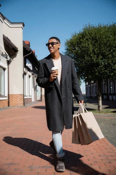 Longitud completa de hombre afroamericano en abrigo de moda y gafas de sol caminando con bebida para llevar y bolsas de compras - foto de stock