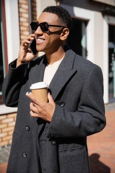 Hombre afroamericano de moda sonriendo durante la conversación en el teléfono inteligente y la celebración de la bebida para llevar en la calle urbana - foto de stock