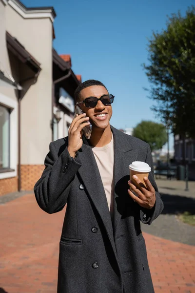 Homem americano africano alegre no casaco elegante e óculos de sol falando no celular enquanto segurando copo de papel na rua — Fotografia de Stock