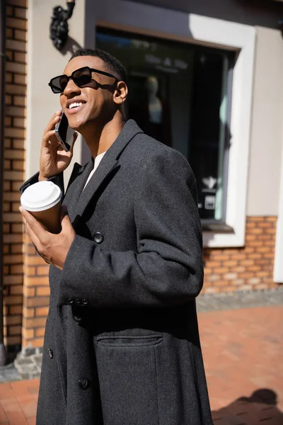 Alegre afroamericano hombre en abrigo y gafas de sol celebración de café para ir y hablar en el teléfono inteligente en la calle - foto de stock