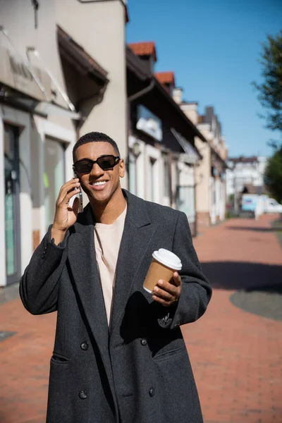 Homme afro-américain souriant et élégant avec du café pour aller parler sur un téléphone mobile près des bâtiments flous — Photo de stock
