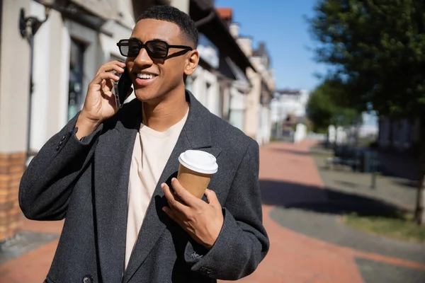 Hombre afroamericano feliz en gafas de sol y abrigo hablando en el teléfono inteligente y sosteniendo taza de papel en la calle - foto de stock