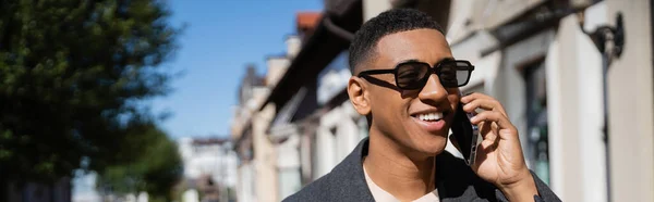 Щасливий афроамериканський чоловік в модних сонцезахисних окулярах розмовляє на смартфоні на розмитій вулиці, банер — стокове фото