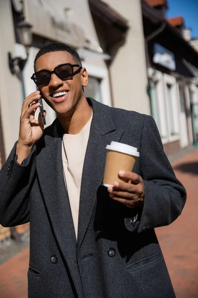 Веселий афроамериканський чоловік в сонцезахисних окулярах і модне пальто тримає паперовий стаканчик і розмовляє на смартфоні на розмитій вулиці — стокове фото