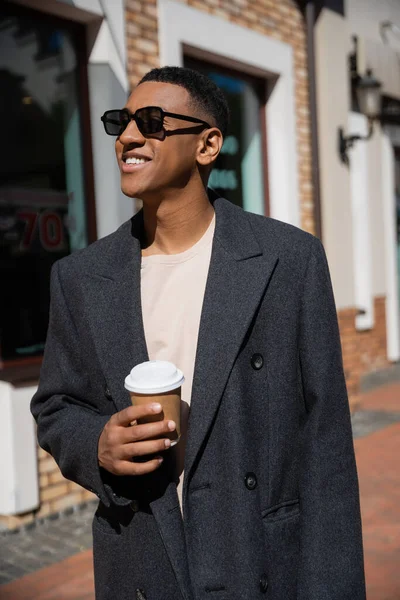 Hombre afroamericano alegre y de moda en gafas de sol de pie con taza de papel y mirando hacia otro lado - foto de stock