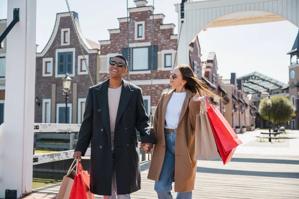 Mujer alegre con bolsas de compras mirando novio afroamericano de moda en el puente - foto de stock
