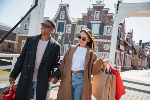 Zufriedenes und stilvolles gemischtrassiges Paar mit Einkaufstaschen Händchen haltend auf der Stadtbrücke — Stockfoto