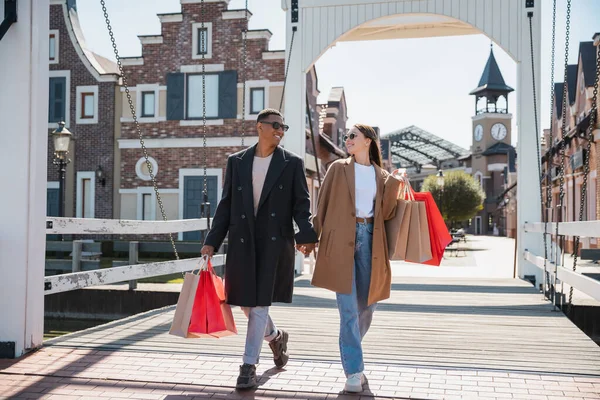 Volle Länge von fröhlichen und trendigen multiethnischen Paar Händchen halten, während zu Fuß auf der Stadtbrücke mit Einkaufstaschen — Stockfoto