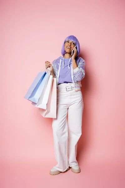 Comprimento total de mulher americana africana alegre em peruca roxa falando no smartphone e segurando sacos de compras em rosa — Fotografia de Stock