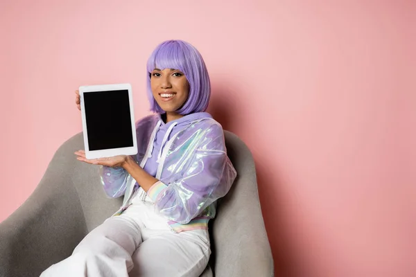 Mulher americana africana satisfeito em peruca roxa segurando tablet digital com tela em branco e sentado em poltrona em rosa — Fotografia de Stock