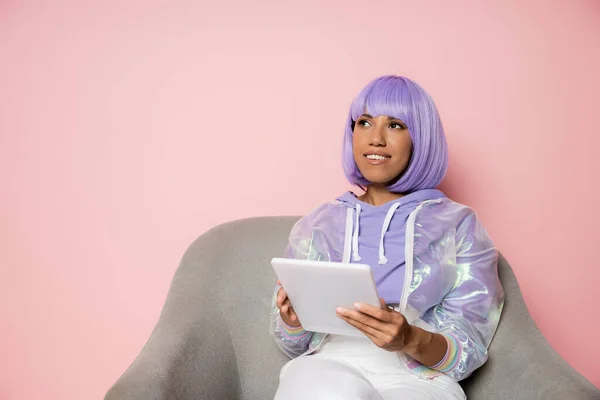 Весела афроамериканка в фіолетовій перуці з використанням цифрового планшета і сидить у кріслі ізольовано на рожевому — стокове фото