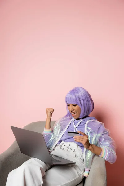 Mujer afroamericana emocionada en peluca púrpura usando el ordenador portátil mientras sostiene la tarjeta de crédito y haciendo compras en línea el viernes negro en rosa - foto de stock