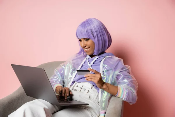 Mulher americana africana alegre em peruca roxa usando laptop enquanto segurando cartão de crédito e fazendo compras on-line na sexta-feira preta em rosa — Fotografia de Stock