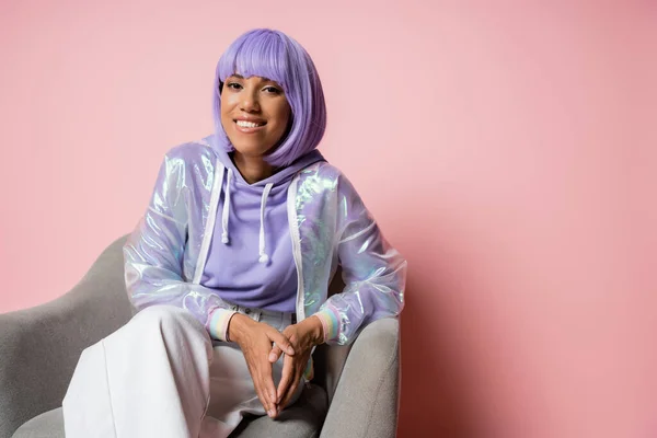 Happy young african american woman in purple wig sitting on grey armchair on pink — Stock Photo