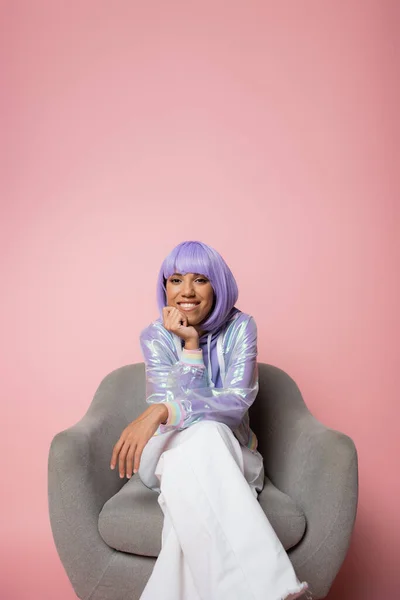 Joyeuse femme afro-américaine en perruque violette souriante assise sur un fauteuil gris sur rose — Photo de stock